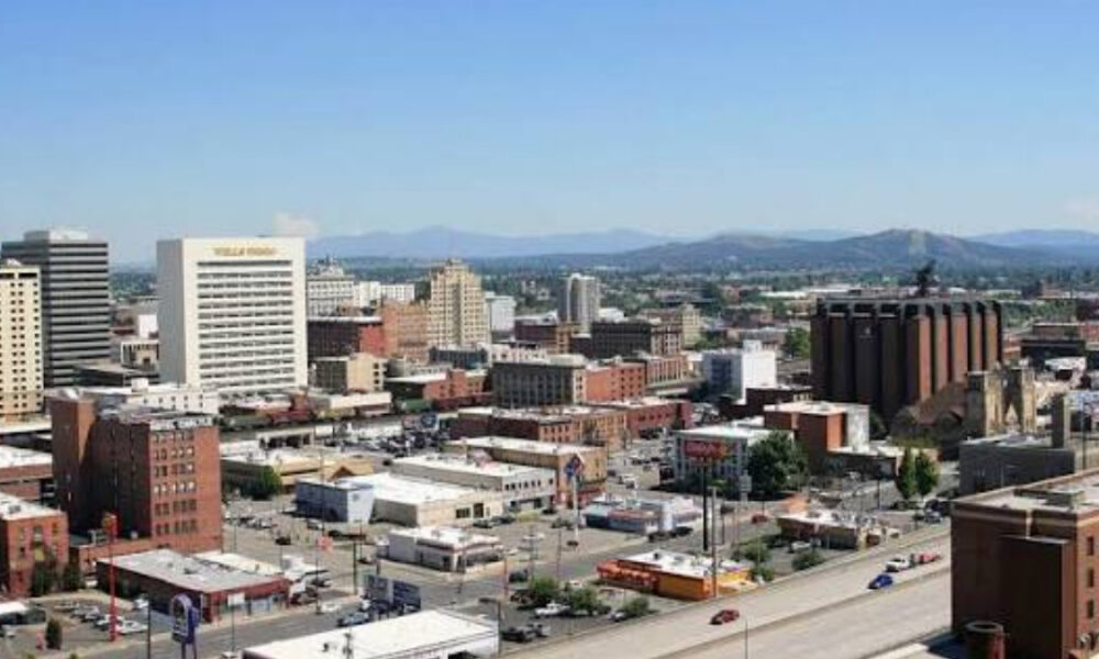 A downtown cityscape with mixed brick and concrete buildings and distant mountains under a clear blue sky.