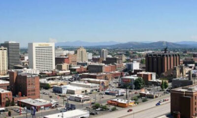 A downtown cityscape with mixed brick and concrete buildings and distant mountains under a clear blue sky.