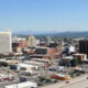 A downtown cityscape with mixed brick and concrete buildings and distant mountains under a clear blue sky.
