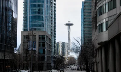 Urban street view with modern glass towers; Space Needle rises between skyscrapers in the distance, cars on the road and leafless trees lining the street.