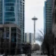 Urban street view with modern glass towers; Space Needle rises between skyscrapers in the distance, cars on the road and leafless trees lining the street.