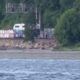 Freight train cars along a track beside a rocky riverbank, with a person in a red jacket near the cars and trees in the background