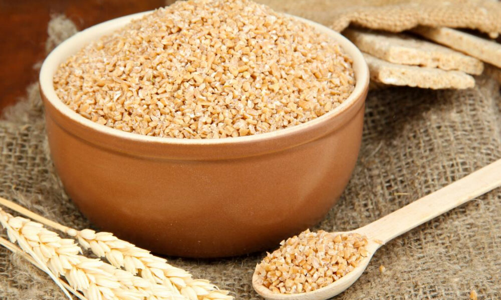 Bowl of bulgur wheat in a ceramic dish with a wooden spoon and wheat stalks on burlap fabric (rustic setting)