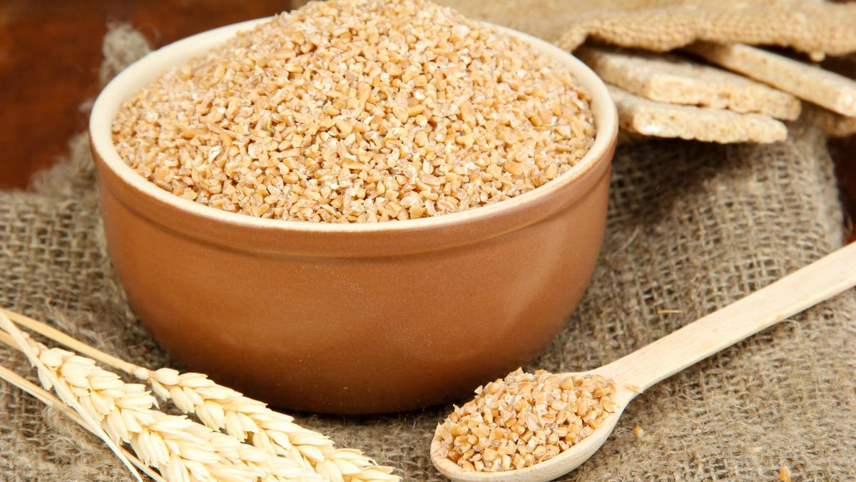 Bowl of bulgur wheat in a ceramic dish with a wooden spoon and wheat stalks on burlap fabric (rustic setting)