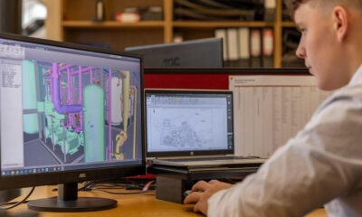 Engineer at a desk editing a 3D CAD model of a mechanical piping system on a monitor without cluttered background