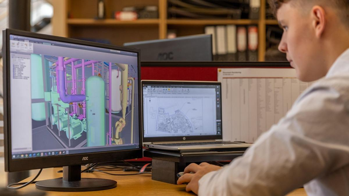 Engineer at a desk editing a 3D CAD model of a mechanical piping system on a monitor without cluttered background