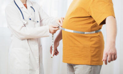 Doctor measures a patient's waist with a tape measure during a medical exam on a light background.
