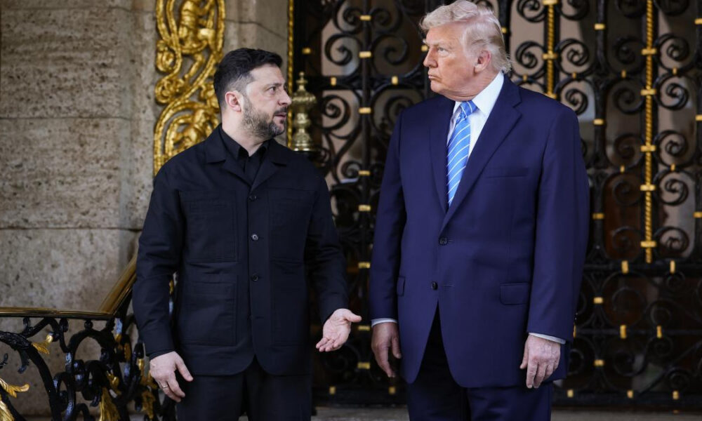 Two men in formal attire stand on a stone staircase in front of an ornate black and gold gate, chatting.