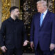 Two men in formal attire stand on a stone staircase in front of an ornate black and gold gate, chatting.