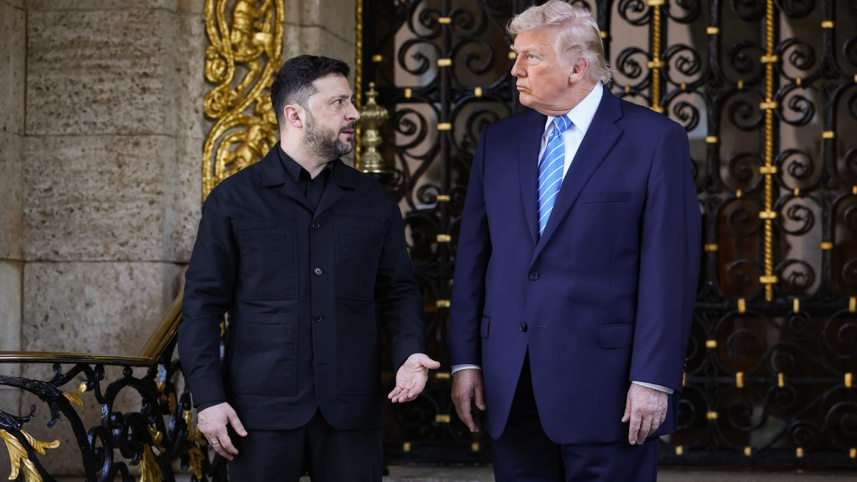 Two men in formal attire stand on a stone staircase in front of an ornate black and gold gate, chatting.