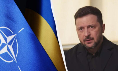 Blue NATO flag on the left and a man in a dark suit speaking indoors on the right, separated by a curved white divider.