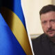 Blue NATO flag on the left and a man in a dark suit speaking indoors on the right, separated by a curved white divider.