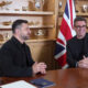 Two men sit at a wooden table in a formal meeting room with Ukrainian and British flags in the background.
