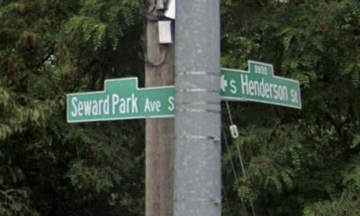 Intersection street signs on a pole reading Seward Park Ave S and Henderson St with leafy trees in the background.