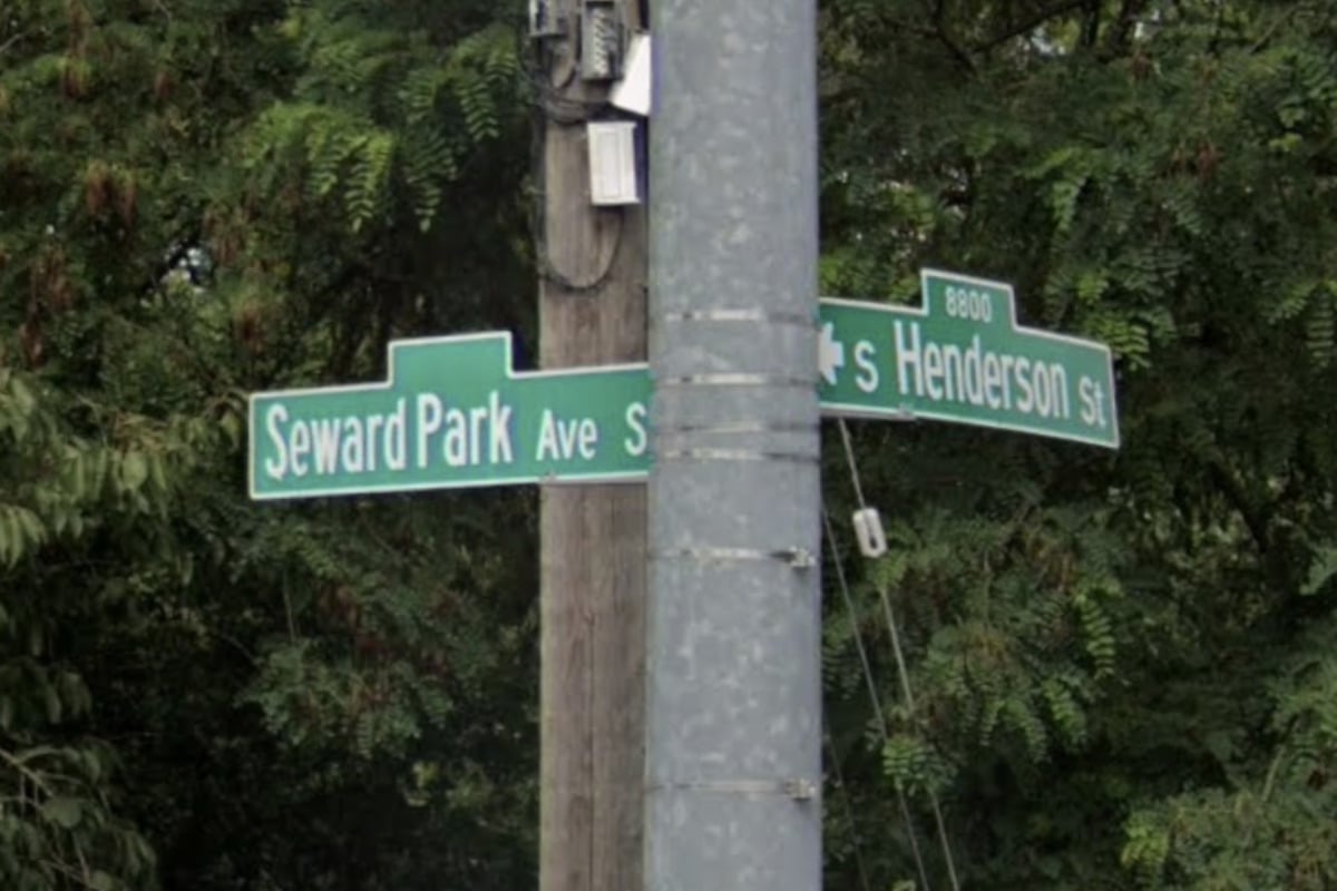 Intersection street signs on a pole reading Seward Park Ave S and Henderson St with leafy trees in the background.