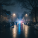 Rainy night on a residential street; two pedestrians cross the wet road with umbrellas as blue police lights glow in the distance.