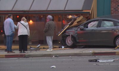 Car crashed into a storefront, glass shattered and debris on the sidewalk, bystanders nearby inspecting the scene