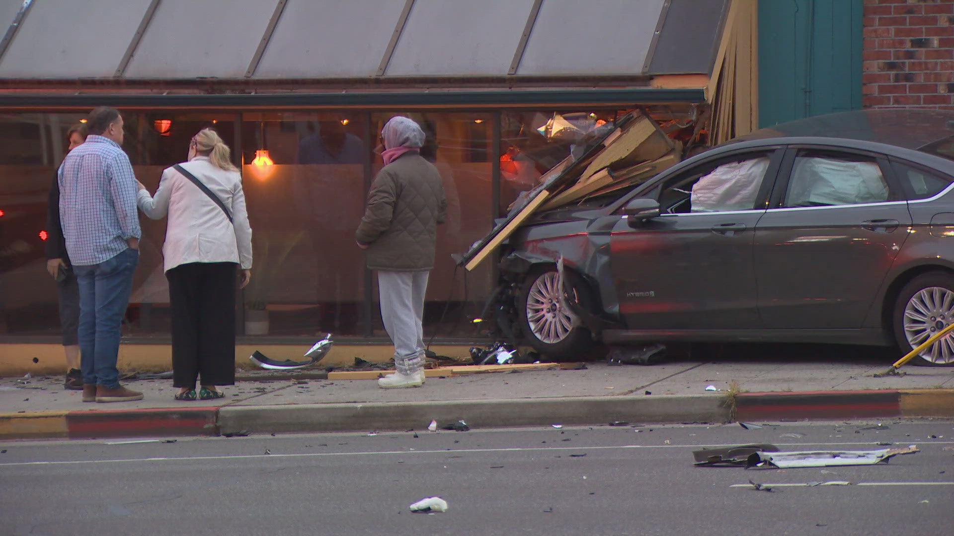 Car crashed into a storefront, glass shattered and debris on the sidewalk, bystanders nearby inspecting the scene