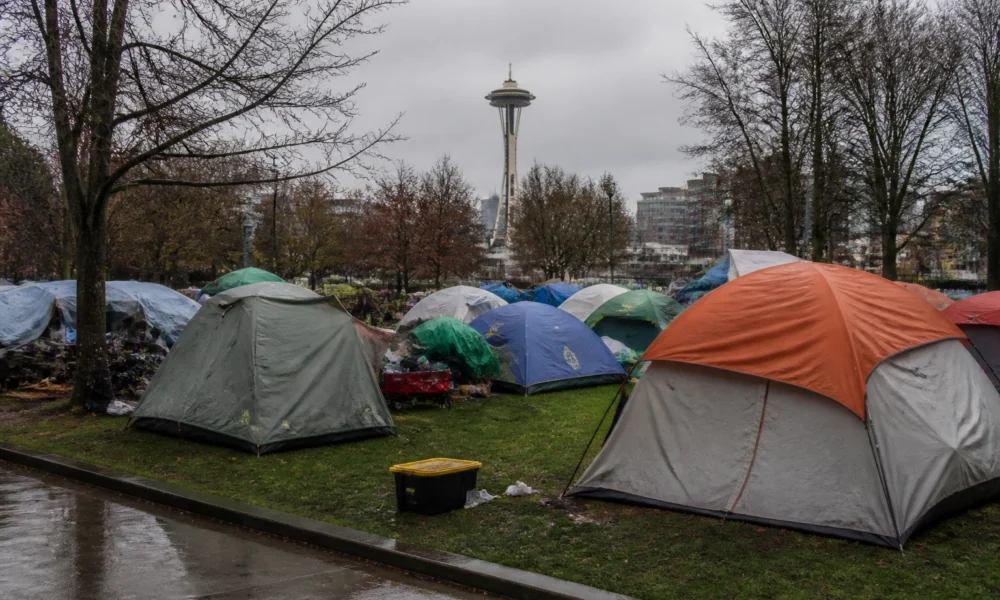 Tents pitched in a city park with the Space Needle visible in the background on a cloudy day, bare trees surrounding the scene and wet pavement nearby.