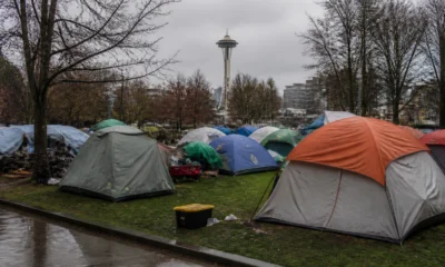 Tents pitched in a city park with the Space Needle visible in the background on a cloudy day, bare trees surrounding the scene and wet pavement nearby.