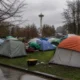 Tents pitched in a city park with the Space Needle visible in the background on a cloudy day, bare trees surrounding the scene and wet pavement nearby.