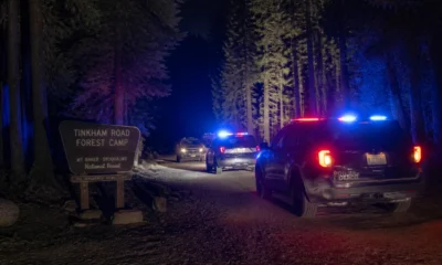 Police cars with flashing red and blue lights on a forest road at night, near a sign for Tinkham Road Forest Camp in Mt. Baker–Snoqualmie National Forest.