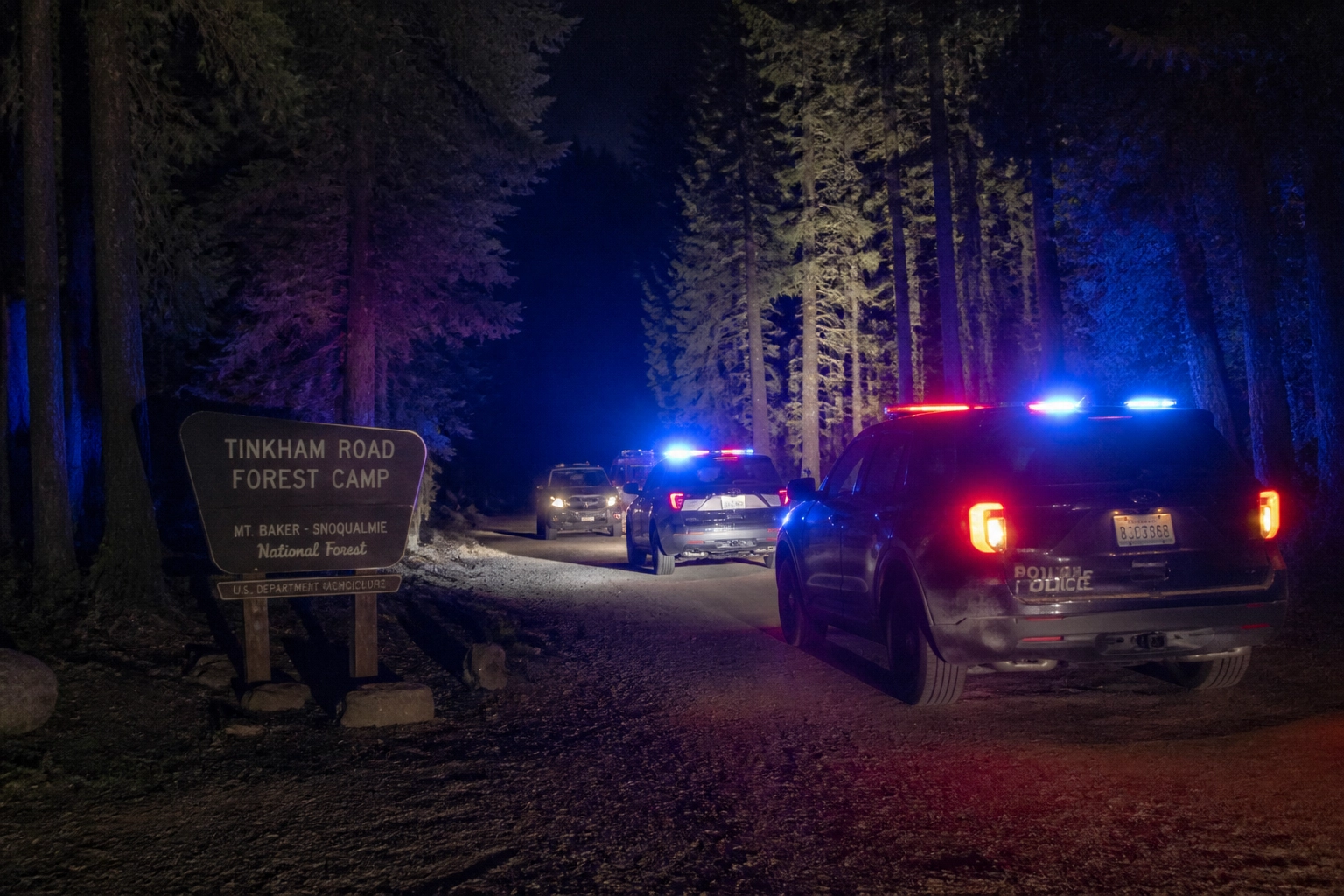 Police cars with flashing red and blue lights on a forest road at night, near a sign for Tinkham Road Forest Camp in Mt. Baker–Snoqualmie National Forest.