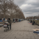 Person with a shopping cart walking along a tents-lined park path under cloudy skies