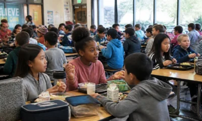 Group of students eating lunch at long cafeteria tables in a bright school dining hall.