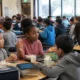 Group of students eating lunch at long cafeteria tables in a bright school dining hall.