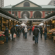 Bustling outdoor market with stalls of fruits and vegetables and shoppers in jackets on a wet cobblestone street.