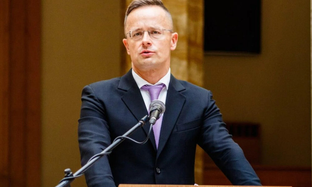 Man in a dark suit and purple tie speaking at a wooden podium with a microphone.
