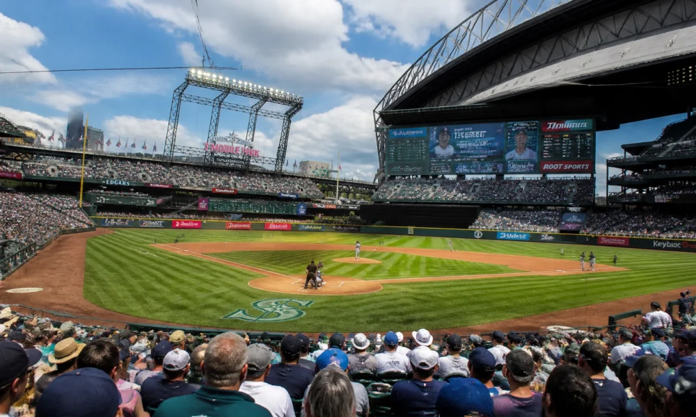 Full view of a baseball stadium with players on the diamond and a large scoreboard above the stands on a sunny day