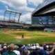 Full view of a baseball stadium with players on the diamond and a large scoreboard above the stands on a sunny day