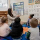 Teacher reads a illustrated story to a circle of young students on a colorful carpet in a bright classroom.