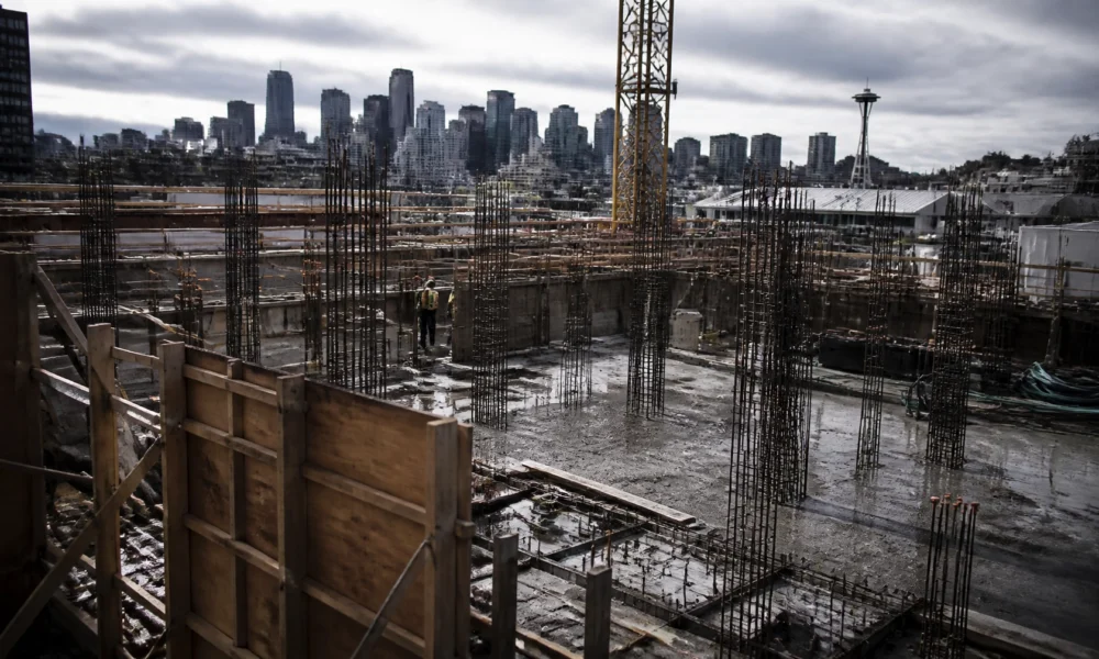 Construction site with exposed steel rebar, wooden formwork, and a worker in a safety vest; Seattle skyline with the Space Needle in the distance.