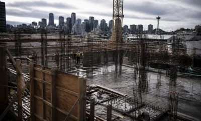Construction site with exposed steel rebar, wooden formwork, and a worker in a safety vest; Seattle skyline with the Space Needle in the distance.