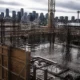 Construction site with exposed steel rebar, wooden formwork, and a worker in a safety vest; Seattle skyline with the Space Needle in the distance.