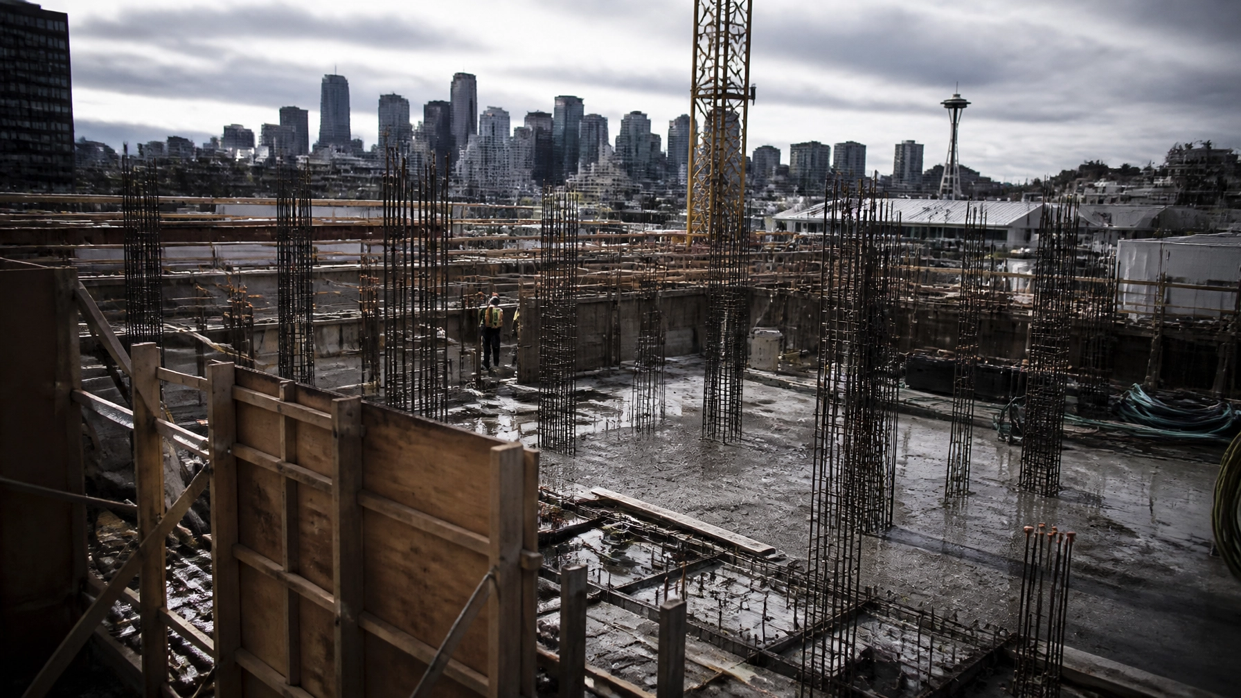 Construction site with exposed steel rebar, wooden formwork, and a worker in a safety vest; Seattle skyline with the Space Needle in the distance.