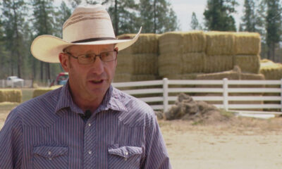 Man wearing a straw cowboy hat and glasses speaks outdoors in a farmyard, with hay bales and a white fence in the background.