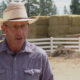Man wearing a straw cowboy hat and glasses speaks outdoors in a farmyard, with hay bales and a white fence in the background.