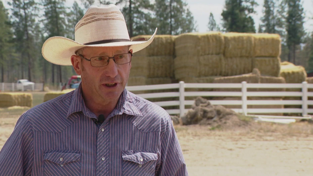 Man wearing a straw cowboy hat and glasses speaks outdoors in a farmyard, with hay bales and a white fence in the background.