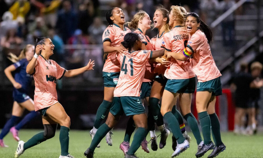 Women’s soccer team in peach uniforms celebrate a goal on the field, arms raised and smiling.