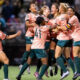 Women’s soccer team in peach uniforms celebrate a goal on the field, arms raised and smiling.