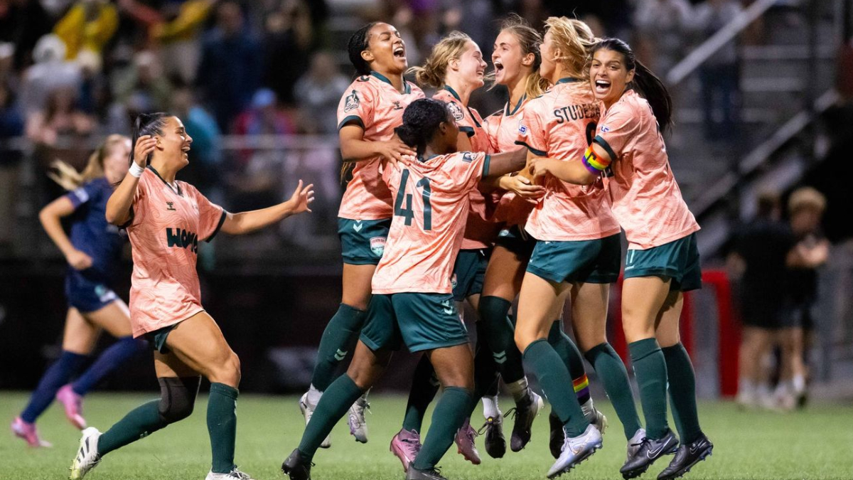 Women’s soccer team in peach uniforms celebrate a goal on the field, arms raised and smiling.