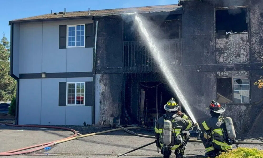Two firefighters spray water on a charred two-story building after a fire.