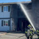 Two firefighters spray water on a charred two-story building after a fire.