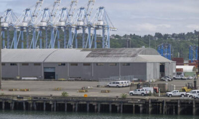Industrial port scene with large gray warehouse, blue gantry cranes, and a row of white service vehicles along the dock edge by the water.