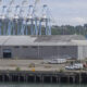 Industrial port scene with large gray warehouse, blue gantry cranes, and a row of white service vehicles along the dock edge by the water.