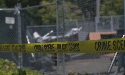 Yellow crime scene tape reading 'CRIME SCENE - DO NOT CROSS' spans a chain-link fence with a blurred outdoor background behind it.